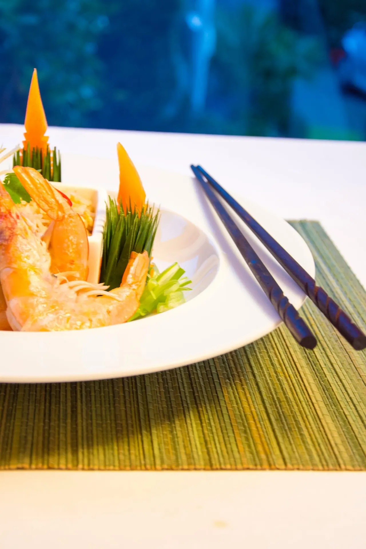 Elegantly plated prawn dish with carved orange garnishes and green onion shoots, served with chopsticks on a woven placemat.