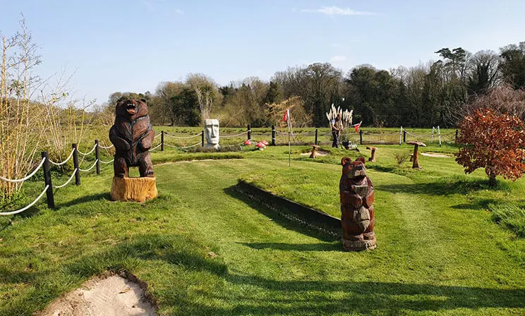 Wooden bear carvings and obstacles scattered across a grass mini golf course with trees in the background.