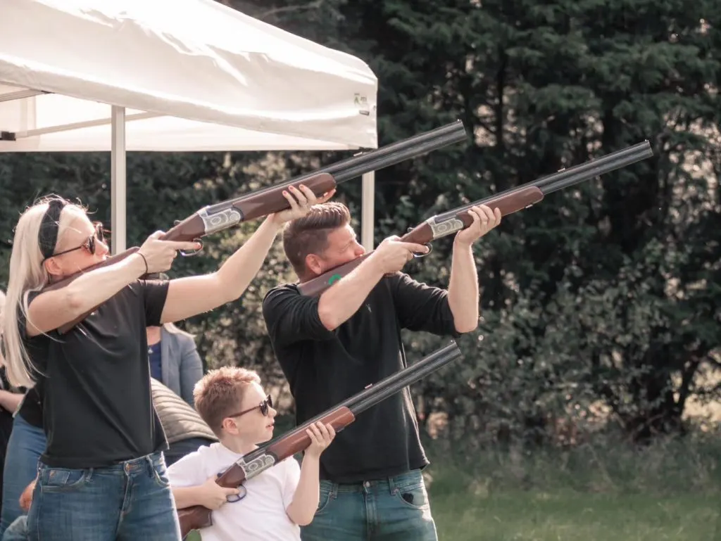 Three people of different ages holding clay pigeon shooting guns under a white canopy, with woodland in the background.
