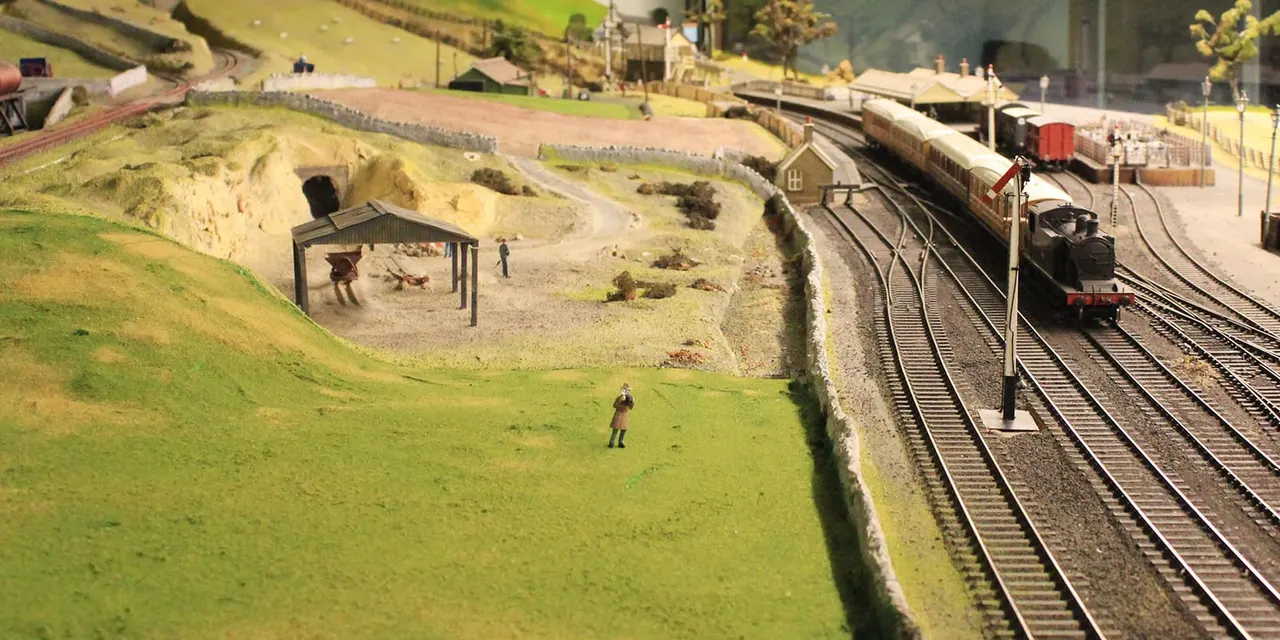 Miniature model railway display with steam locomotive, track, station buildings, and figures on landscaped terrain.