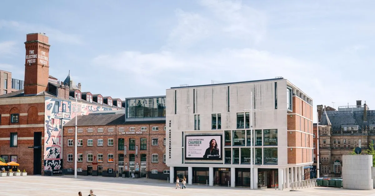 A modern theatre building with glass and stone façade stands beside historic red-brick Victorian structures in an urban public square.