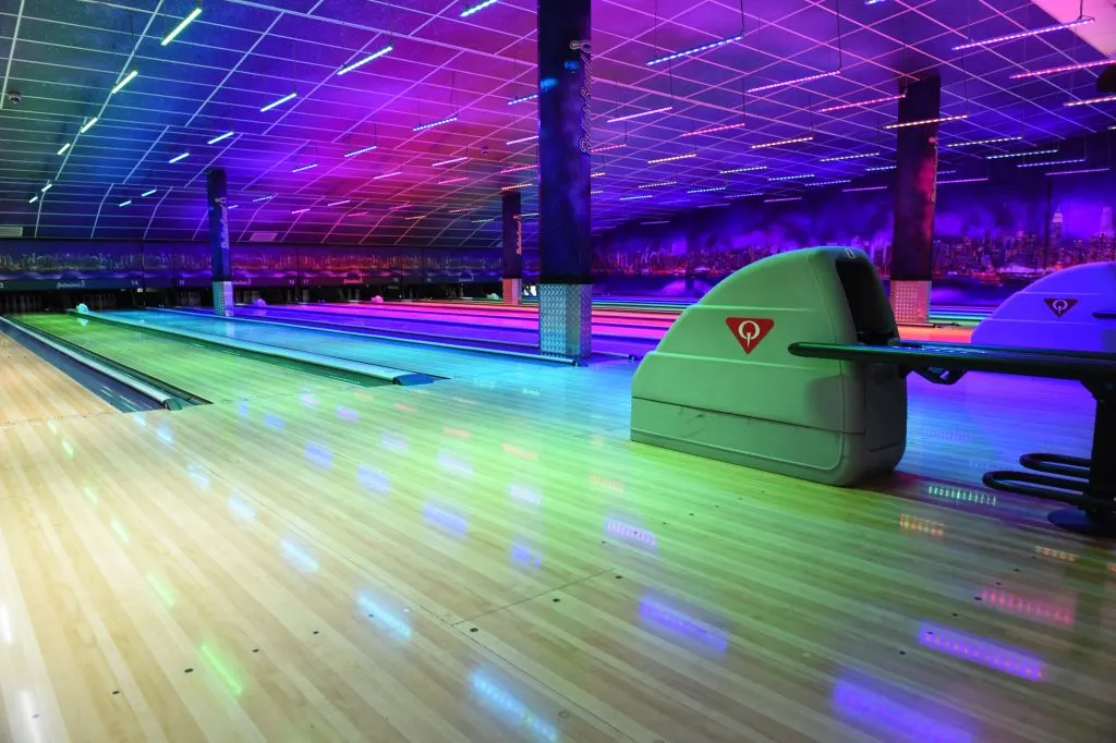 Neon-lit bowling alley with multiple lanes, colourful overhead lights, and pin-setting machines under vibrant purple and blue lighting.