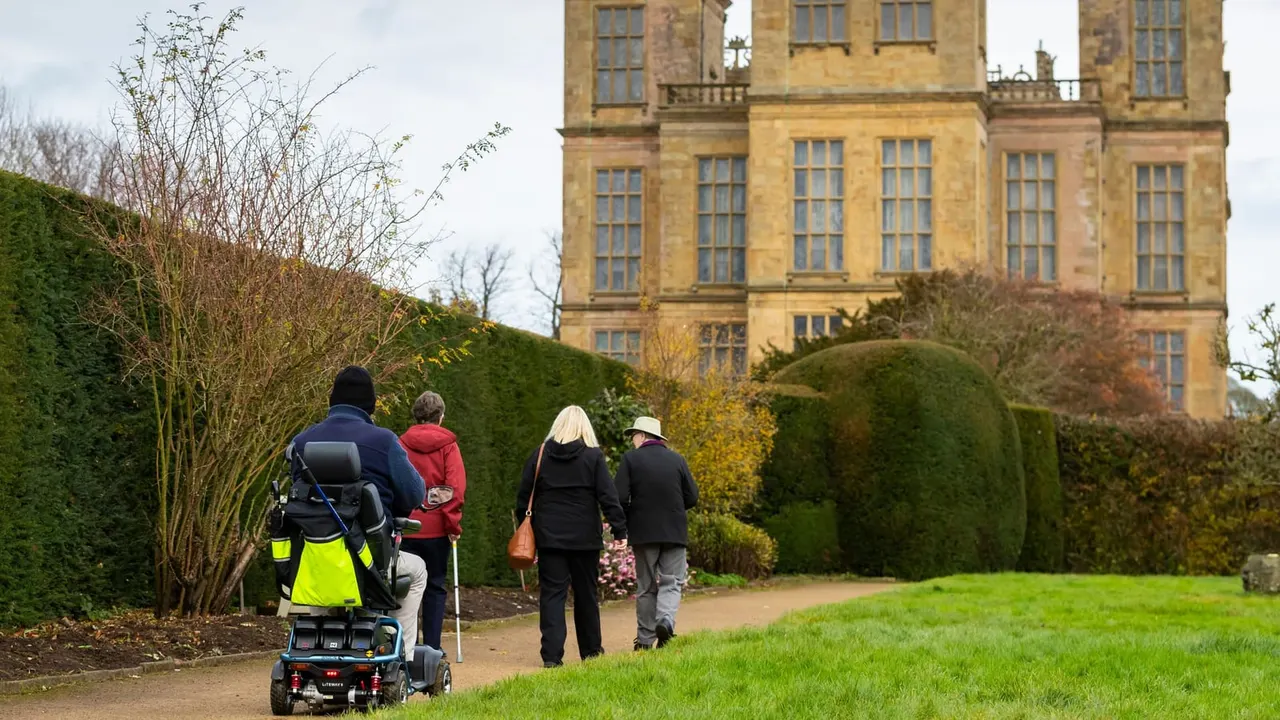Visitors walking and a person with a pushchair on a path through manicured gardens towards a large stone country house.