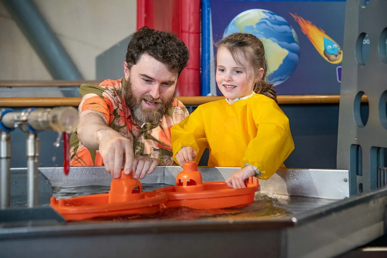 Adult and child playing with a red toy boat in a water trough at an interactive science exhibit.