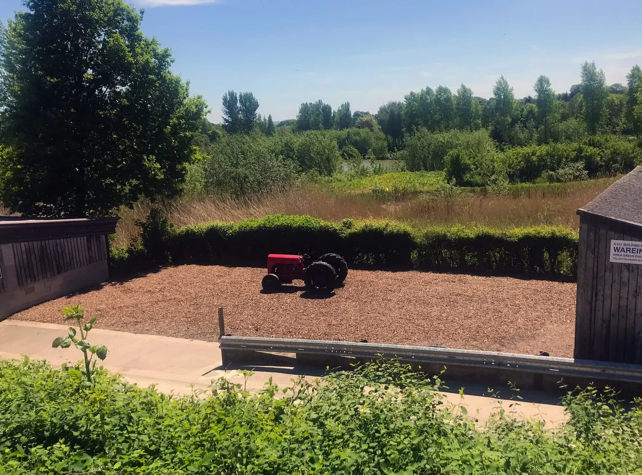 A red vintage tractor sits in a gravel arena surrounded by wooden fencing, with rolling countryside and trees in the background.