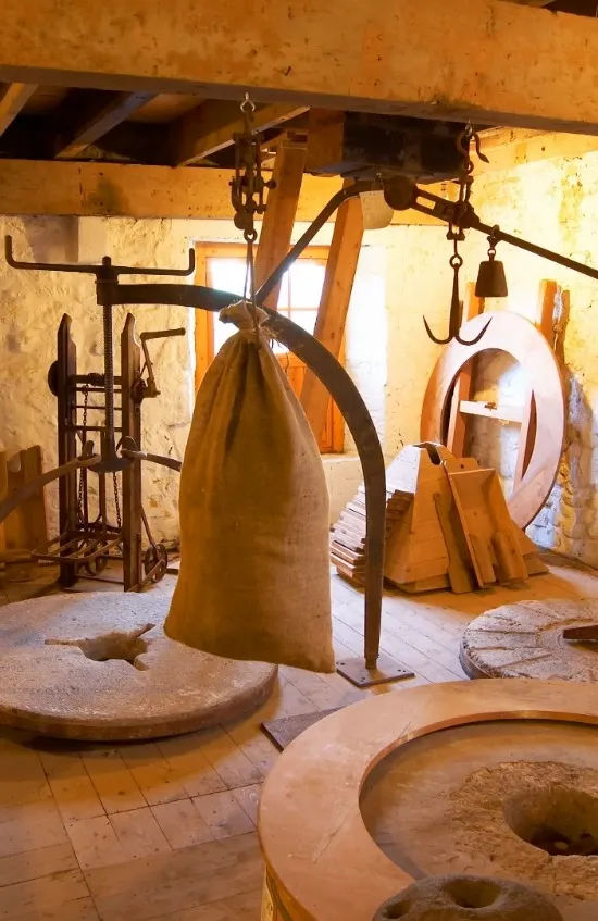 Historic mill interior with large suspended grinding stone and traditional machinery equipment.