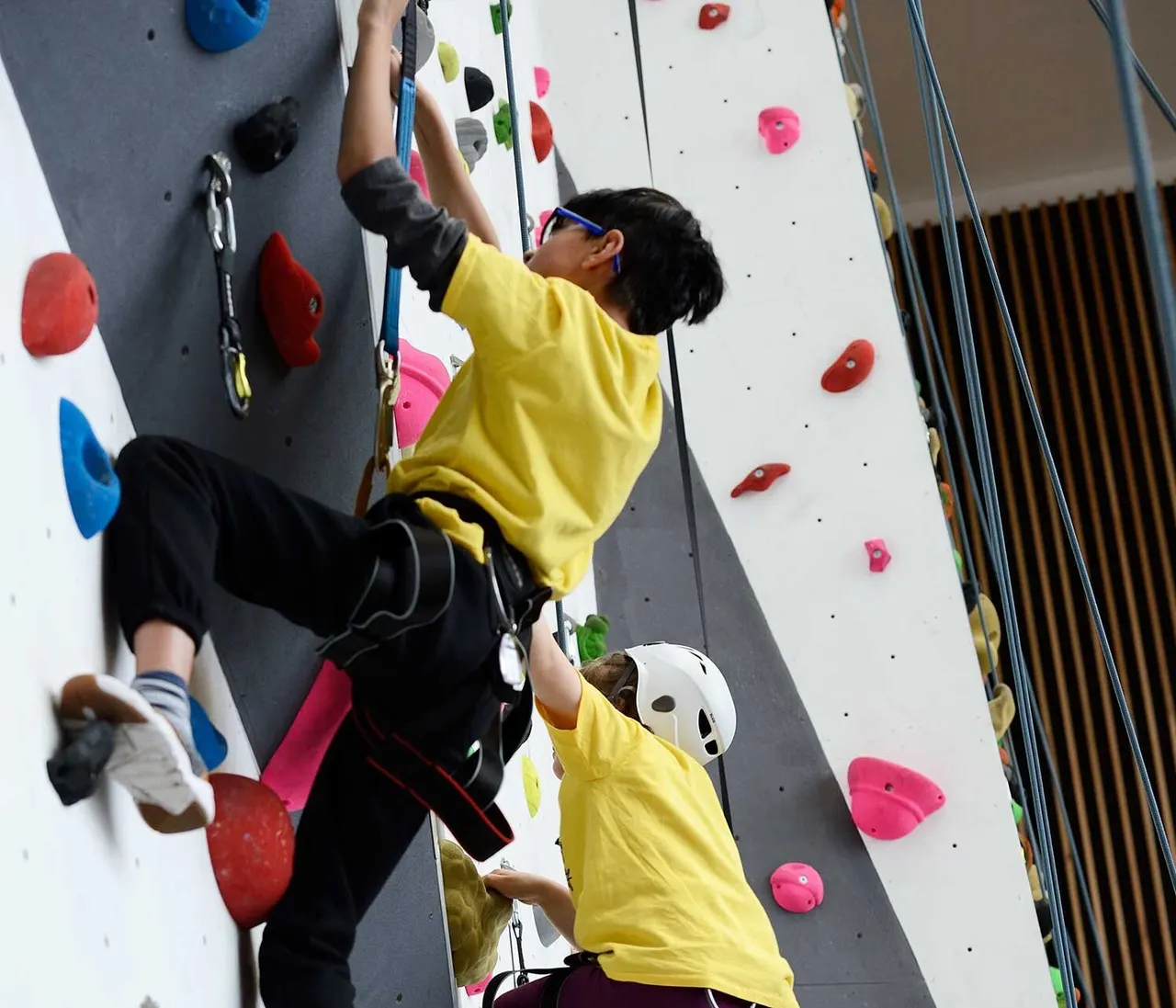 Child in yellow shirt climbing indoor rock wall with coloured holds while wearing safety harness and helmet.