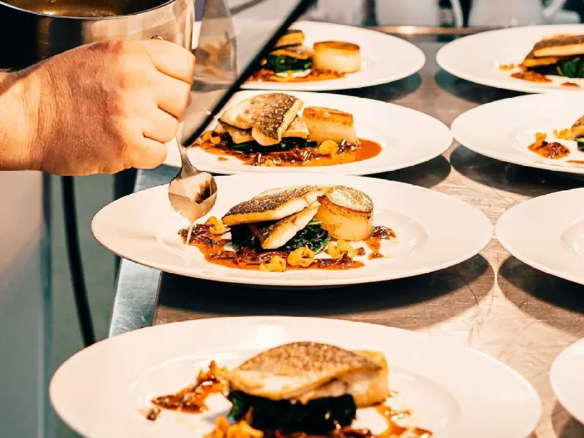 Person holding a glass of wine at a dining table with multiple plated fish dishes garnished with vegetables.
