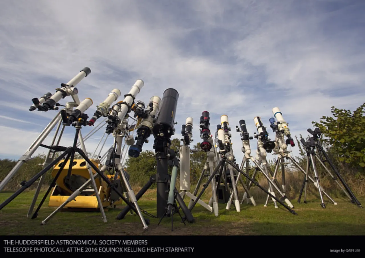 Multiple telescopes mounted on tripods in a field under cloudy sky, with green grass and trees visible.