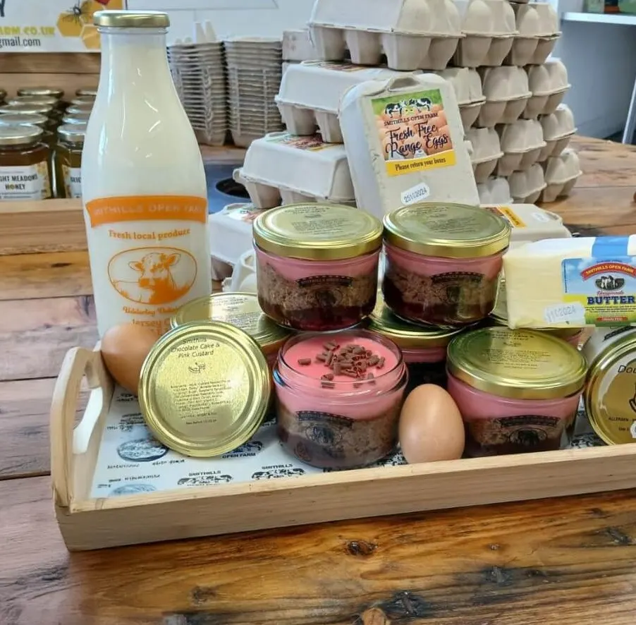 Wooden tray displaying local farm produce including bottled milk, jars of pink and brown layered desserts, eggs, butter and packaged free-range eggs, with egg cartons stacked in background.