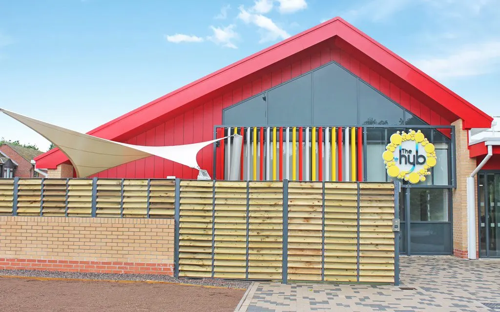 Modern leisure building with distinctive red roof and colourful rainbow-striped window feature, enclosed by wooden fence gate.
