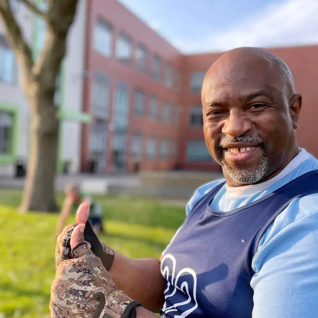 Man giving thumbs up to camera whilst standing on grass in front of a red brick building.