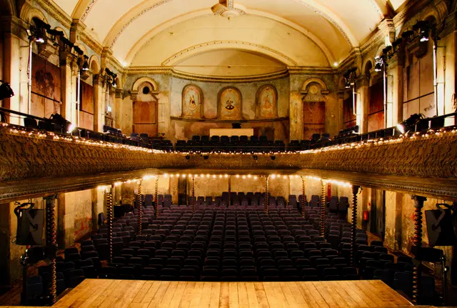 Historic theatre interior with ornate cream and gold arched ceiling, balconies, and rows of dark seats facing a wooden stage.