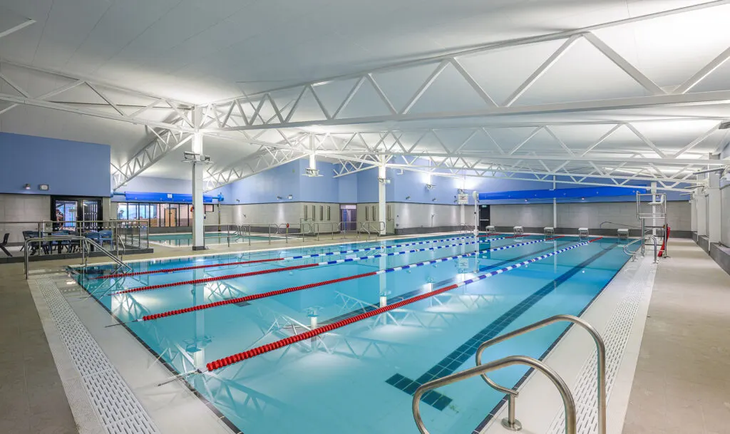 Indoor swimming pool with multiple lanes divided by red lane ropes, viewed from poolside in a modern facility with white steel roof trusses.