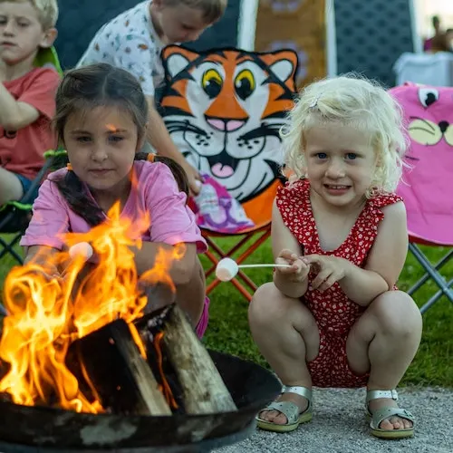 Young children gathered around a small campfire with a tiger mascot decoration behind them.