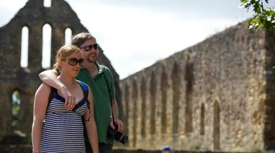 Two visitors in sunglasses stand together viewing the ruins of a medieval stone abbey.