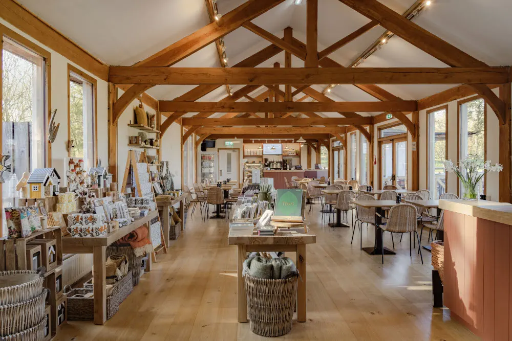 A spacious timber-framed interior with exposed wooden beams, retail displays of homeware and gifts on the left, dining tables and chairs on the right, and large windows overlooking grounds.