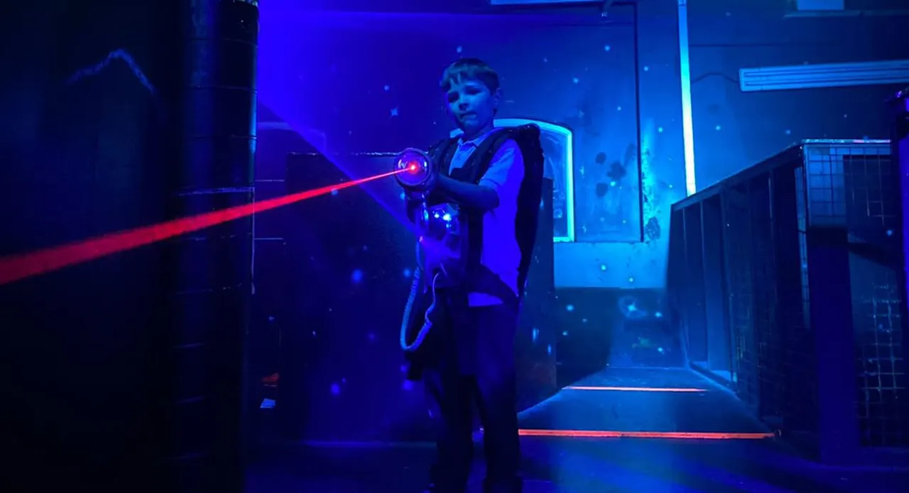 Child aiming a laser gun with red beam in an indoor laser tag arena with blue neon lighting.
