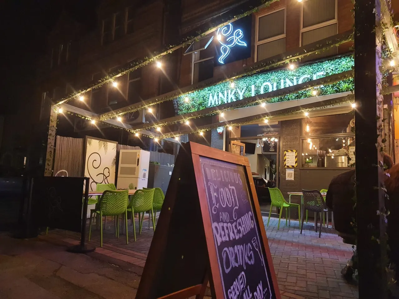 Illuminated outdoor seating area at night with lime green chairs, neon signage, and festoon lighting above a brick building entrance.