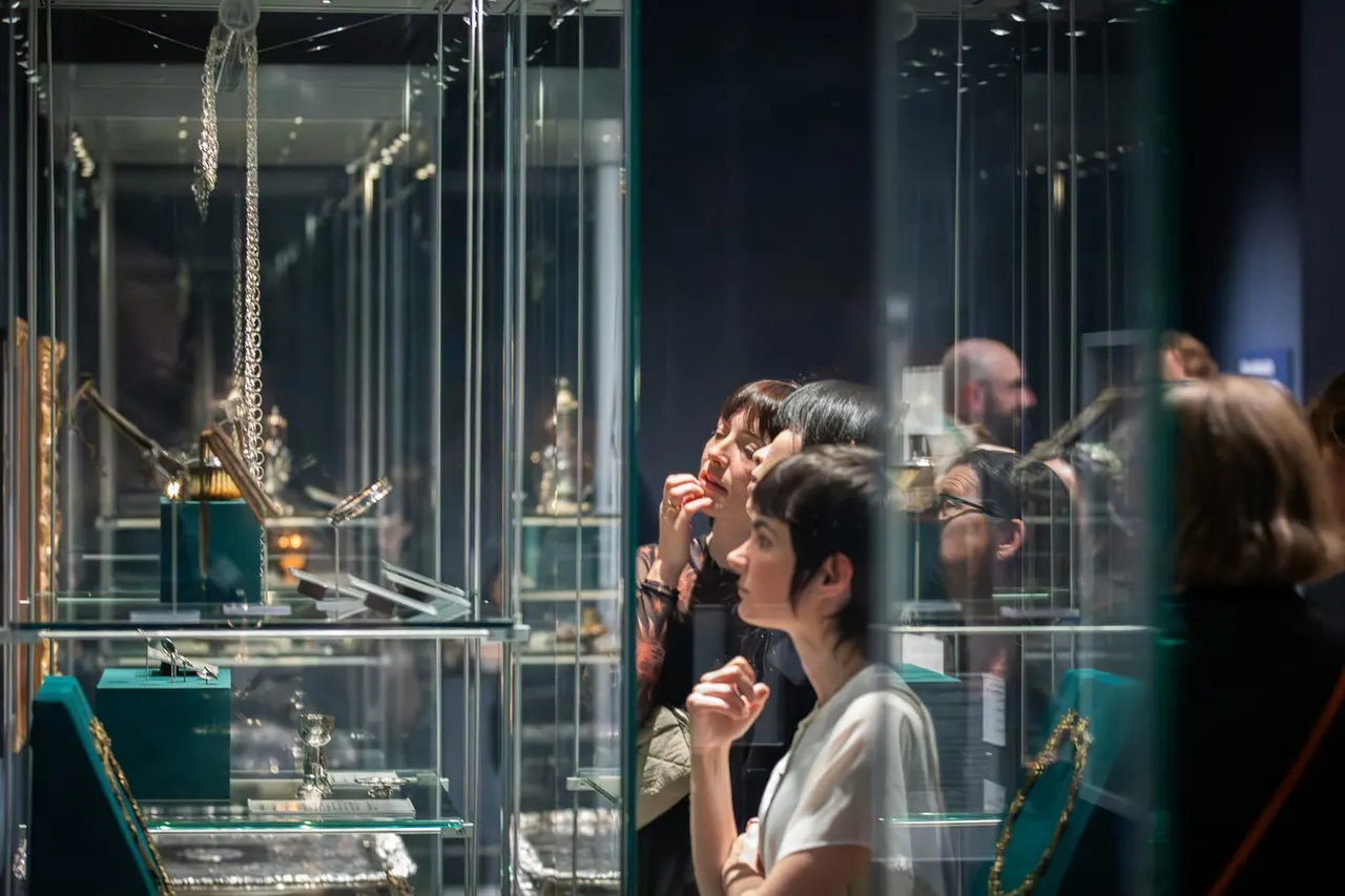 Visitors viewing jewellery and precious objects displayed behind glass cases in a museum gallery.