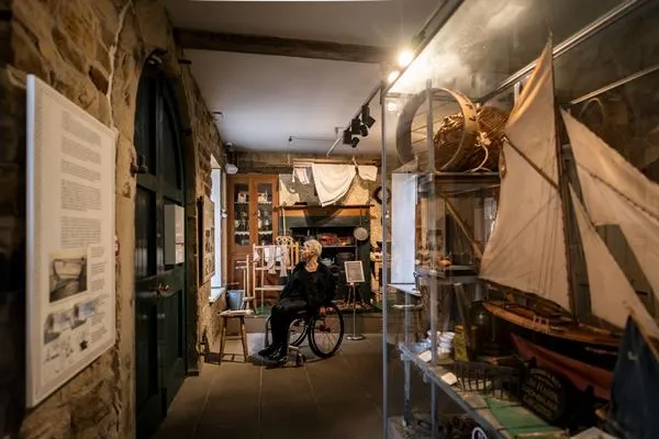 Person in wheelchair exploring museum corridor with maritime artefacts including model sailing ships and informational displays on stone walls.