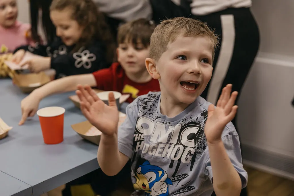 Young child laughing with hands raised at a table during a group activity, with other children and adults nearby.