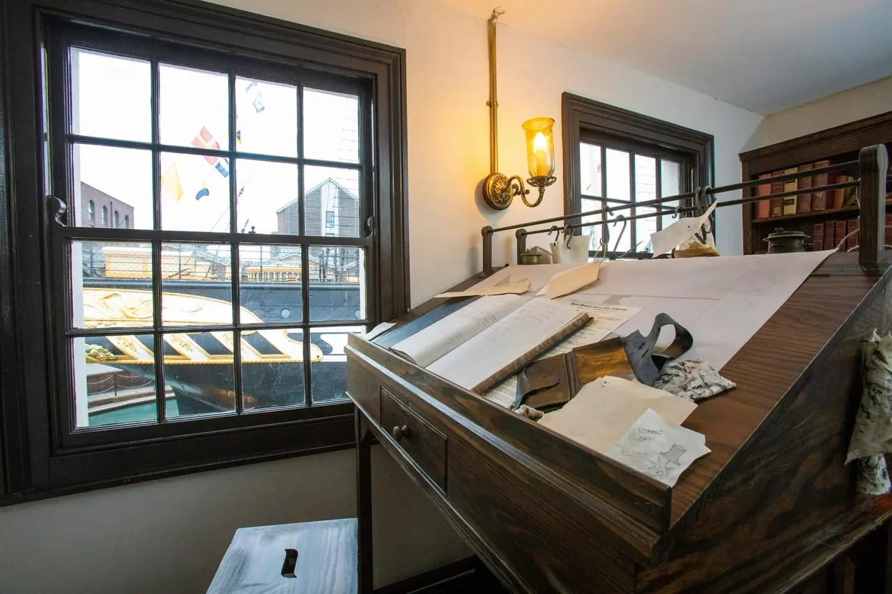 Historic bedroom display with period furniture, blueprints and tools on a desk, overlooking a waterfront through large windows.