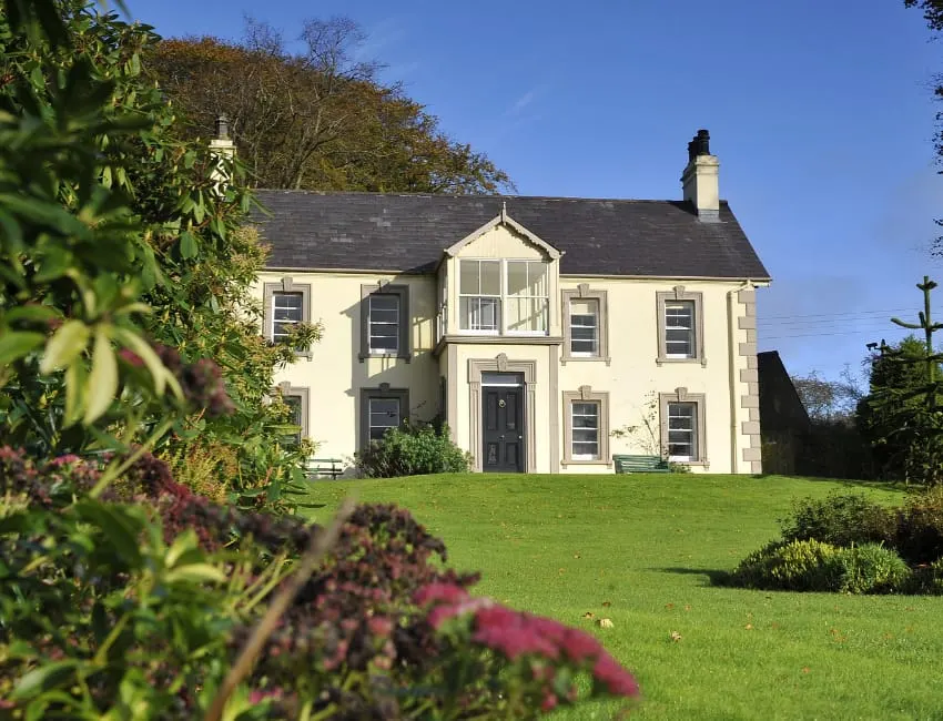Cream-coloured historic house with dark roof set in manicured grounds, flowering shrubs in foreground, mature trees beyond.