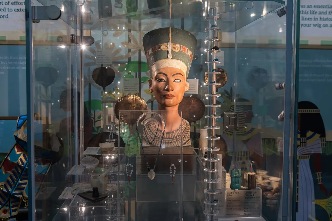 Museum display case containing Egyptian artefacts including a bust sculpture wearing a royal headdress, with jewellery and small objects arranged around it.