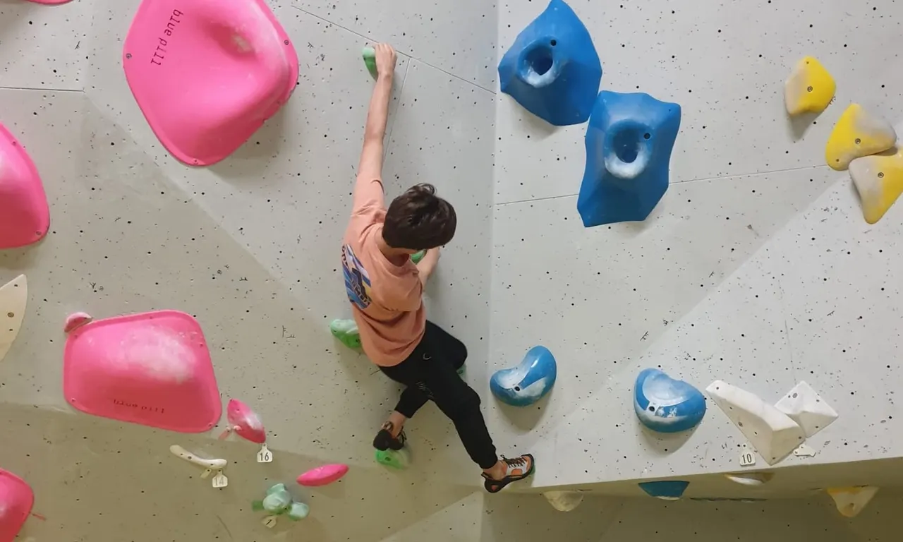 Child climbing an indoor wall with coloured holds, reaching towards pink and blue wall features.