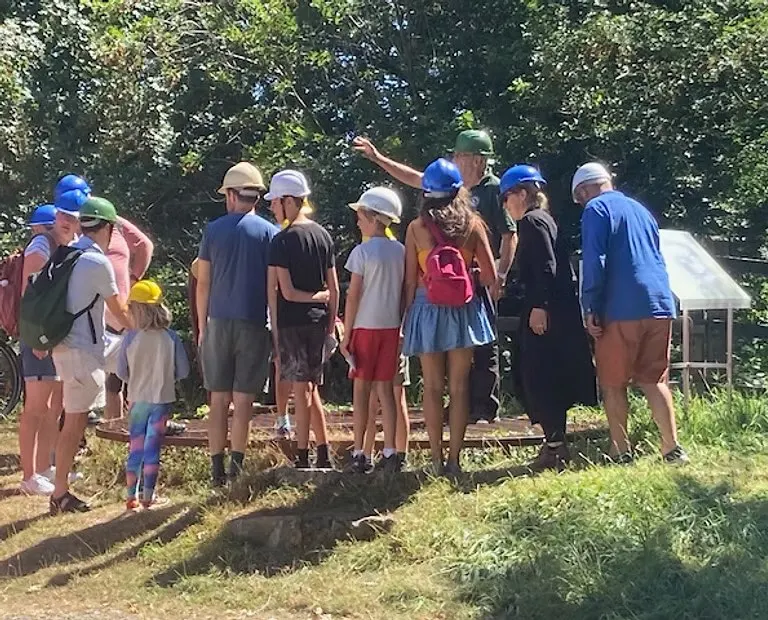 Group of visitors wearing hard hats gathered outdoors at a cave entrance surrounded by trees and vegetation.