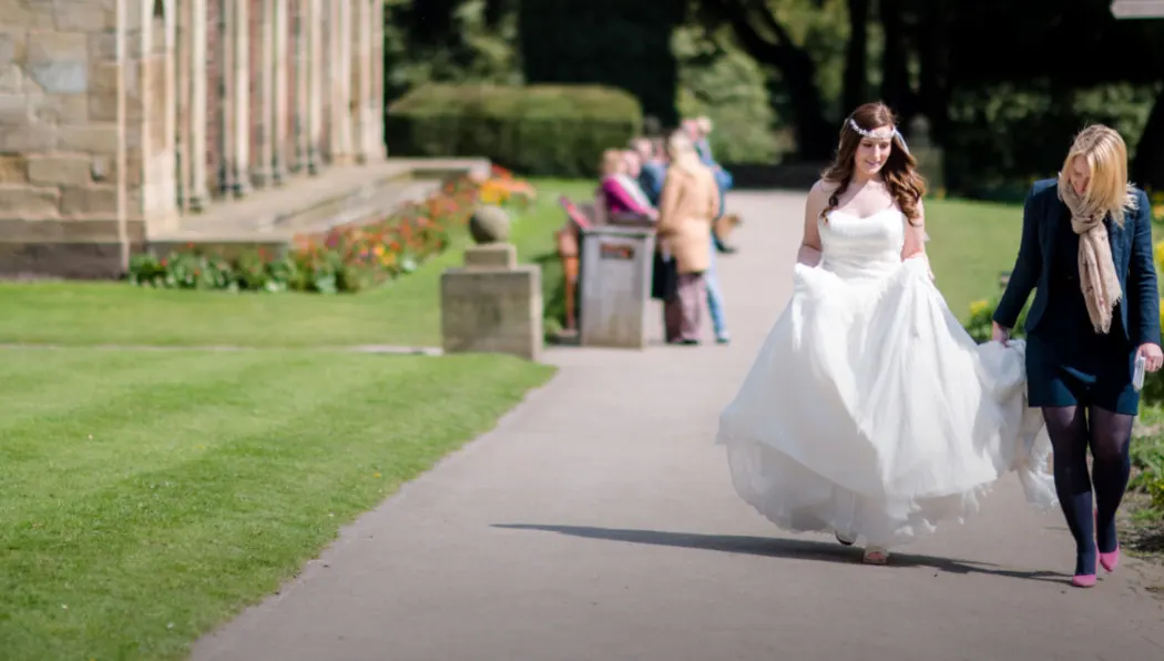 Bride in white wedding dress walking with companion along garden path beside stone building.