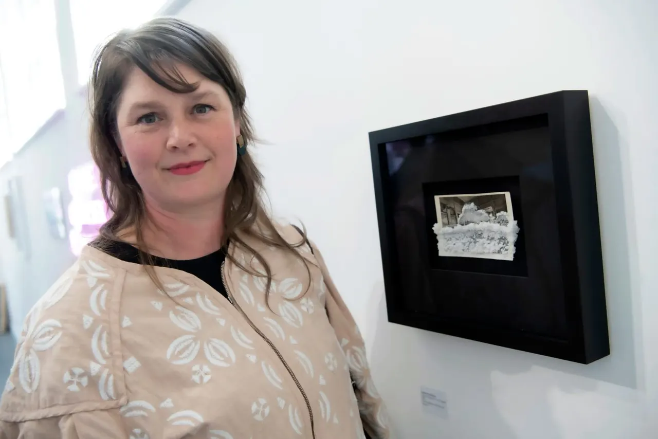 Woman smiling at camera next to framed artwork on white gallery wall.