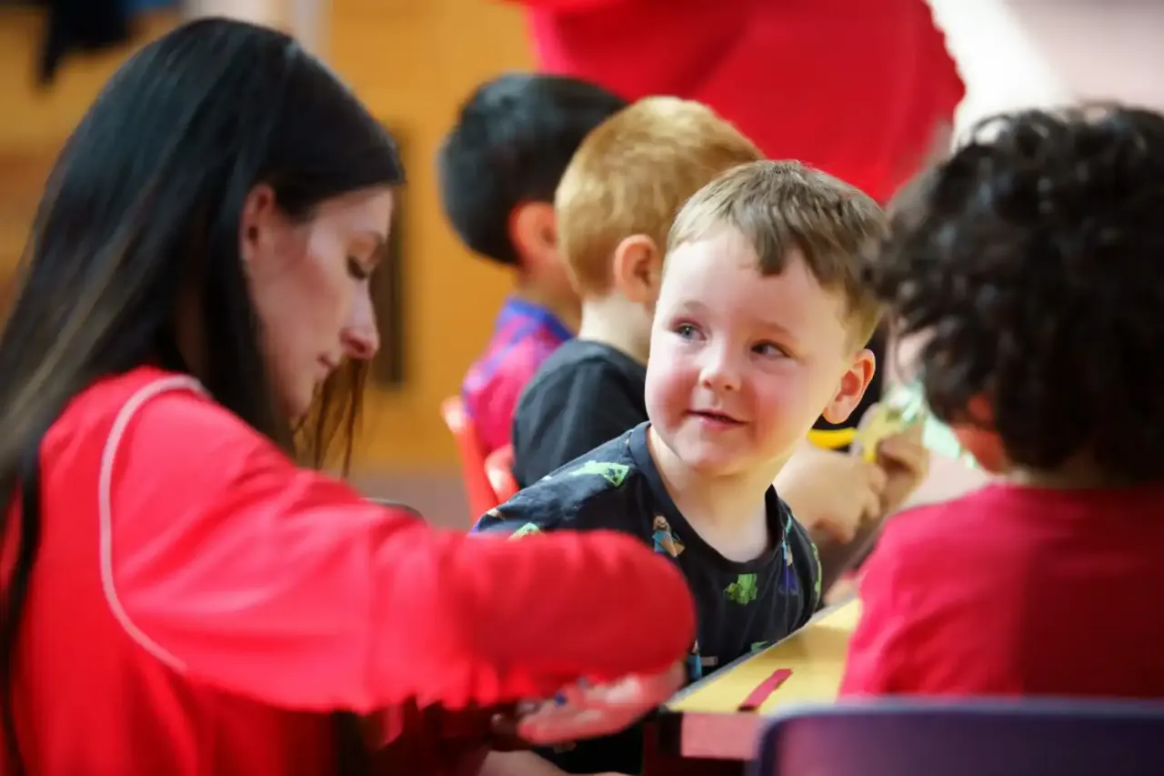 A young child smiling whilst being helped by an adult instructor in red at an indoor sports activity.