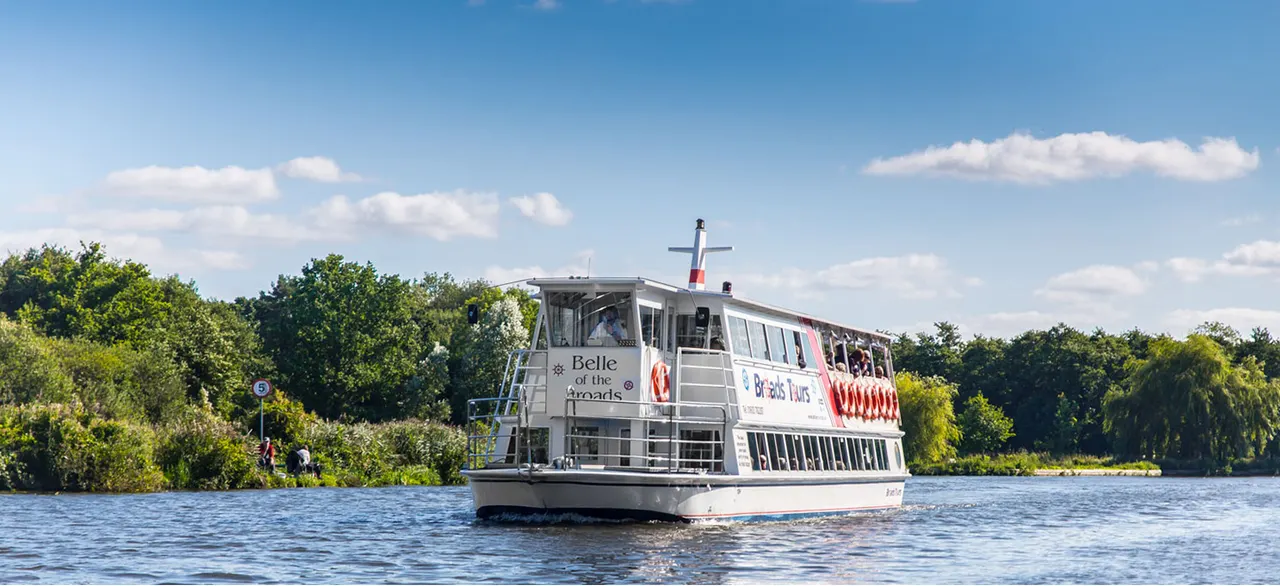 Passenger riverboat cruising on a broad with tree-lined banks under blue sky.
