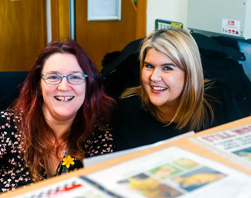 Two smiling women at a reception desk with printed materials displayed in front of them.