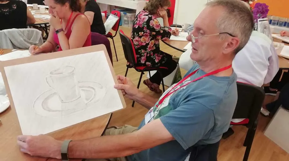 Man holding a pencil sketch of teacups during an art class with other participants seated around tables.