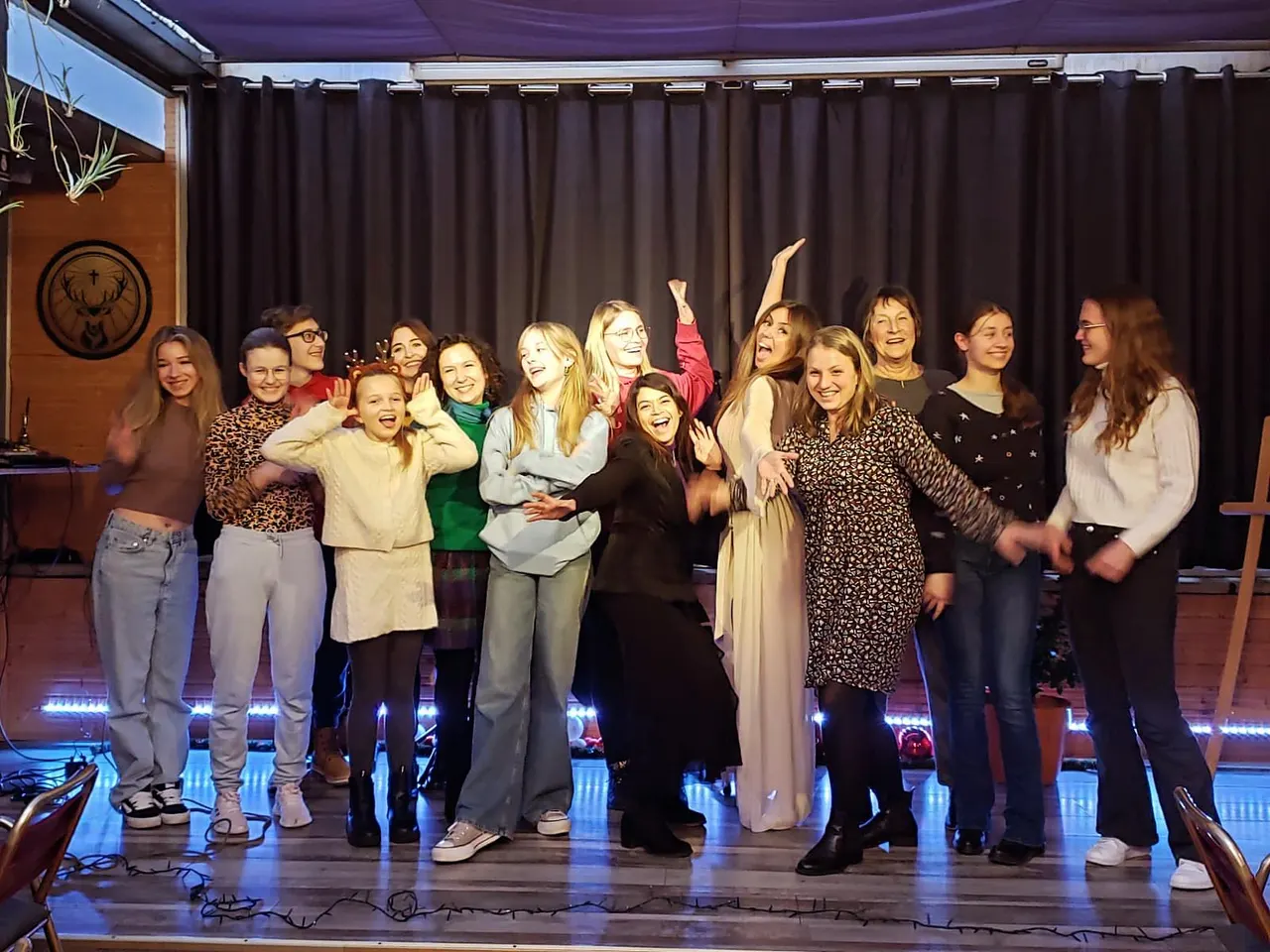 A group of around fifteen women and girls stand together on a lit stage, smiling and posing with raised arms in a celebratory pose.