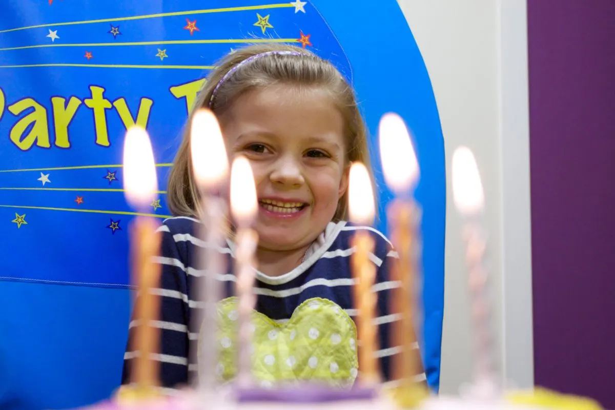 Young child smiling at a birthday cake with lit candles in front of a blue starry backdrop.