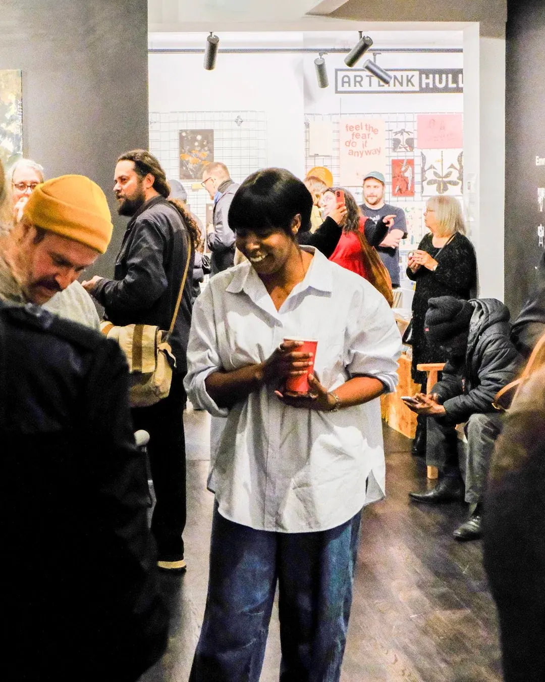 People mingling at an art venue opening or event, with exhibition displays visible on the wall behind them.