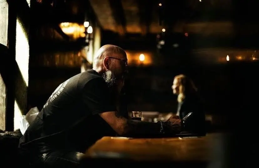 A man with a beard sits at a bar counter in a dimly lit venue, holding a drink.