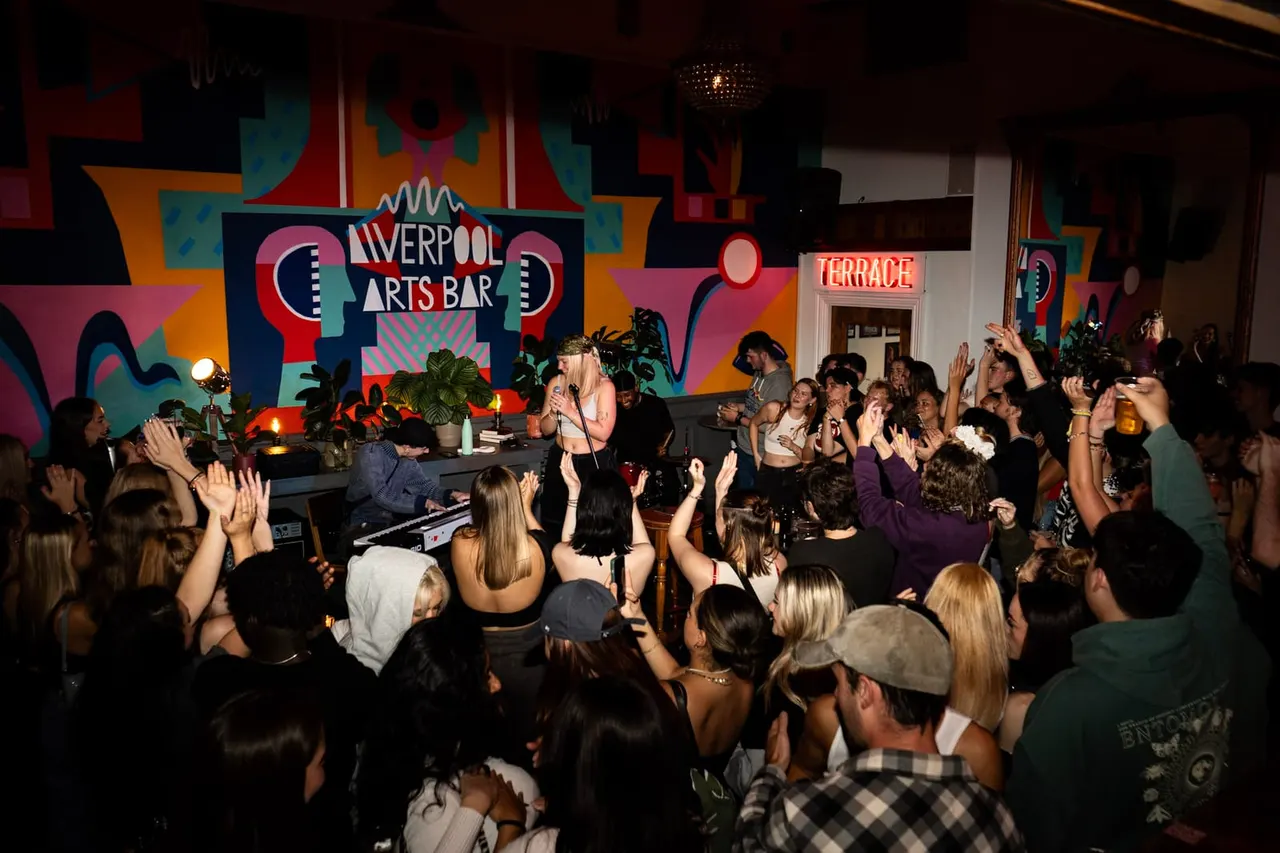 A musician performs on stage to a crowd of dancing and cheering guests in a colourful, vibrant arts bar venue.