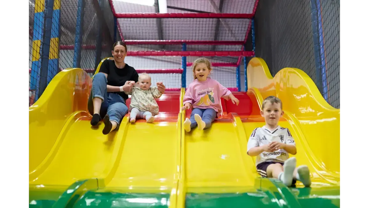 An adult and three young children sliding down bright yellow slides inside an indoor play structure with coloured netting.