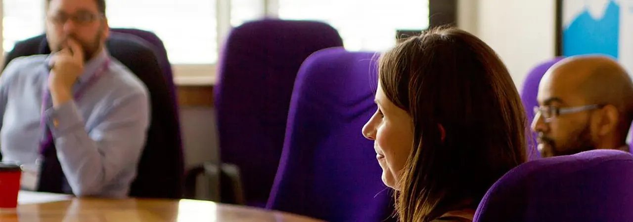 People seated in purple chairs around a table during an indoor meeting or discussion.