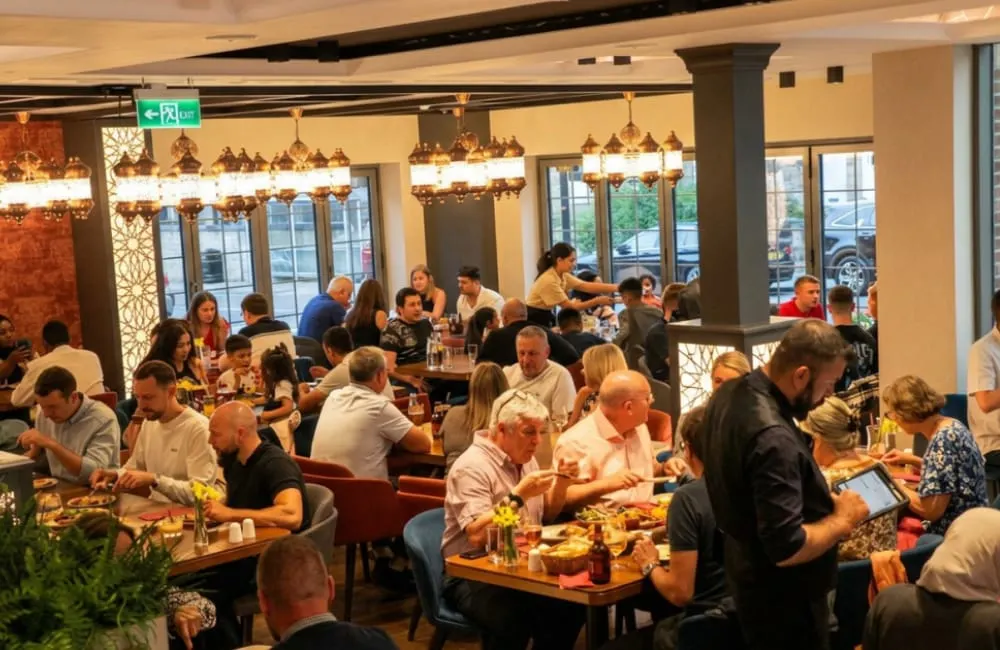 Groups of adults dining and socialising at tables in a modern restaurant with copper pendant lights and floor-to-ceiling windows.