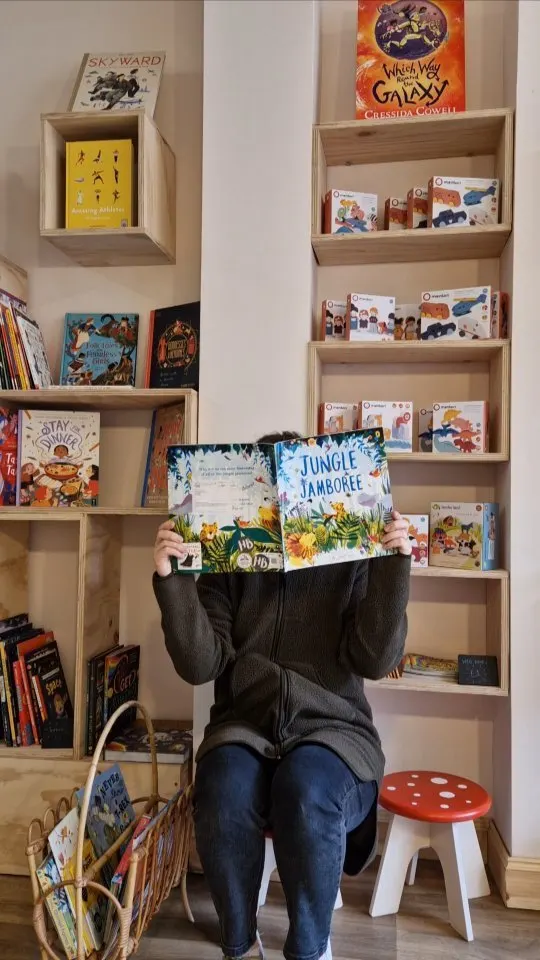 Person sitting in front of wooden shelves holding up a children's picture book titled "Jungle Jamboree".