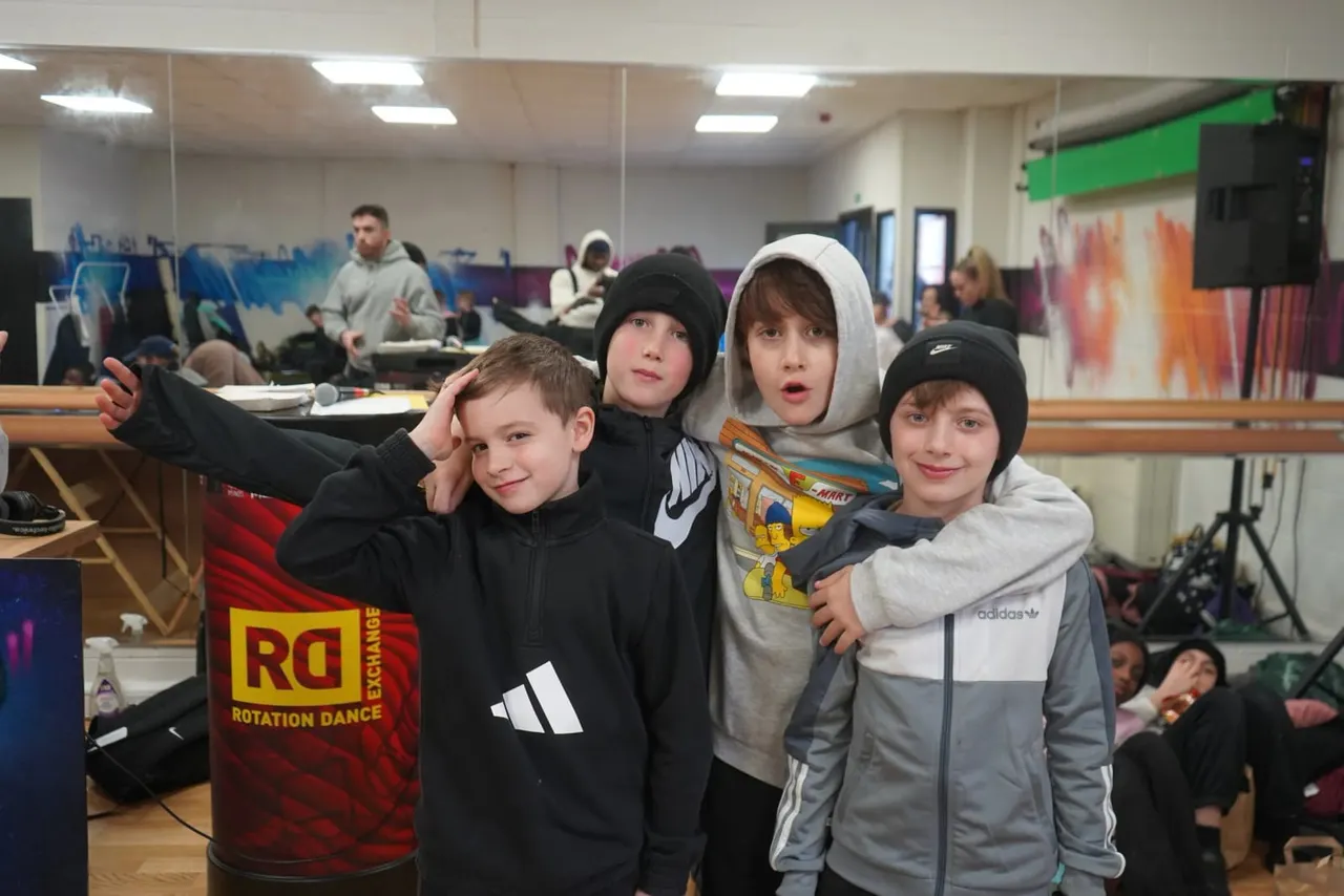 Five children posing together in a dance studio, wearing casual hoodies and beanies, with other people and equipment visible in the background.