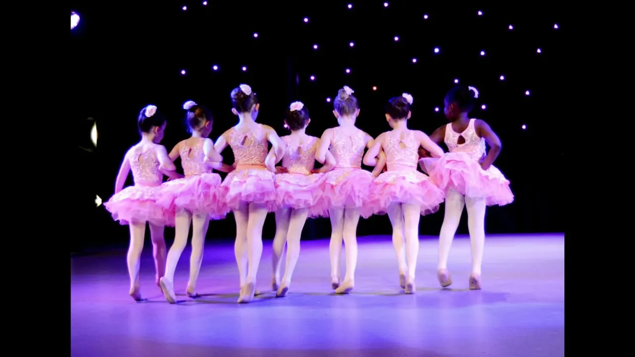 Young ballet dancers in pink tutus performing on stage under purple lighting.