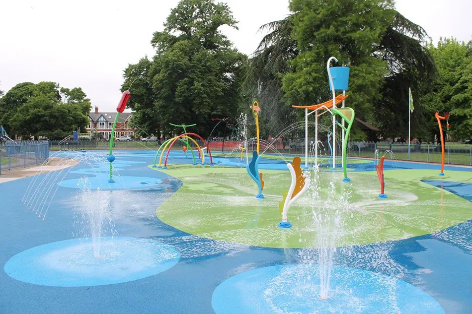 Colourful water play area with fountains, spray jets and climbing structures on blue and green surfacing in a park setting.