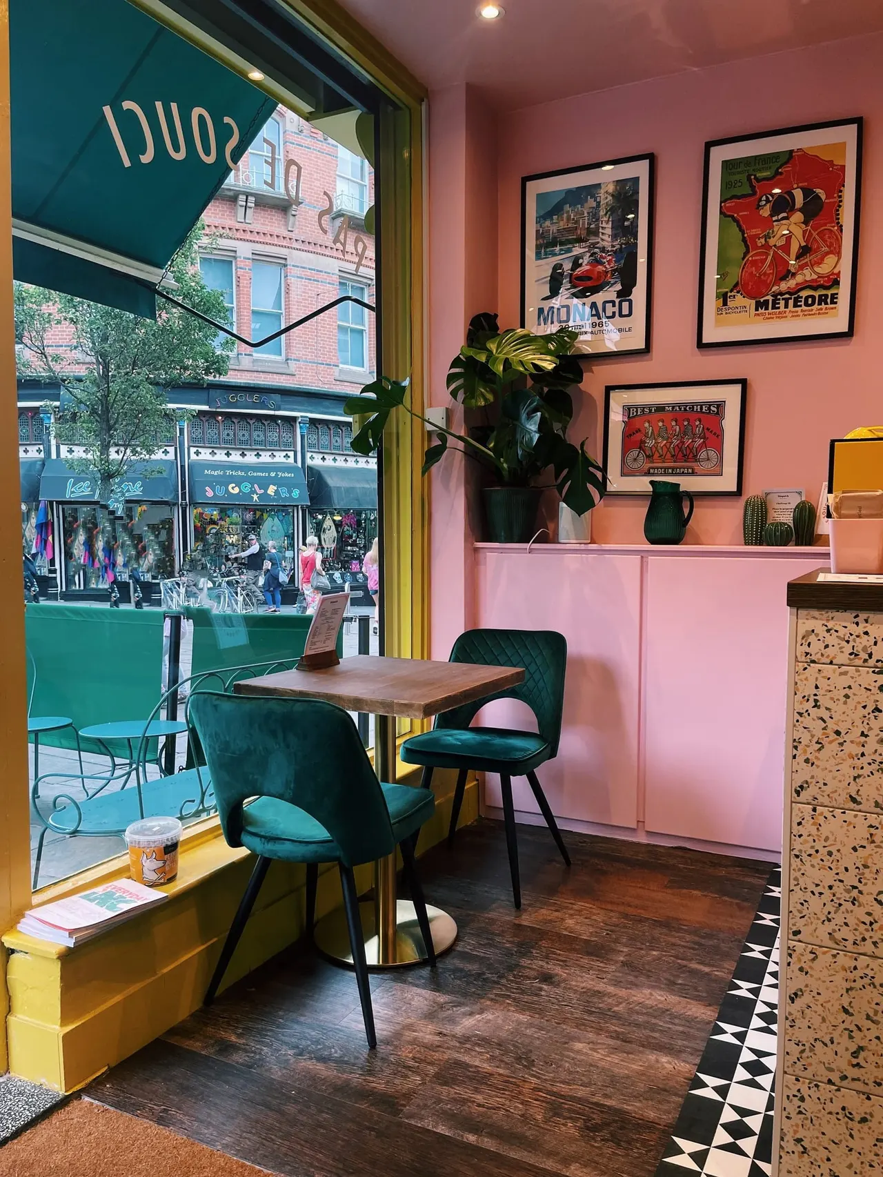 A colourful café interior with teal chairs at a wooden table by a large window, pink walls decorated with vintage posters, potted plants, and a street view outside.